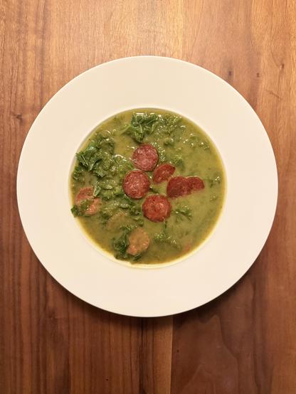 Ein weißer, tiefer Suppenteller. Eine zartgrüne Cremesuppe , darin kräftig dunkelgrüne Streifen von Kohlblättern und paprika-rote Scheiben von gebratener iberischer Wurst.