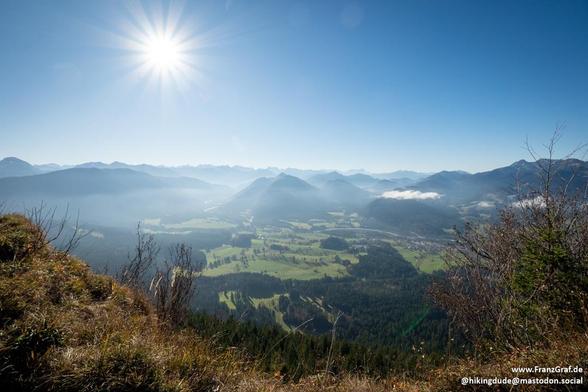 A stunning panoramic view unfolds under a bright, clear sky, where the sun shines brilliantly, casting a warm glow over a picturesque valley. The sun's rays create a starburst effect, adding a magical touch to the scene. Below, a lush valley stretches out, filled with a patchwork of green fields, dense forests, and scattered clusters of buildings, all bathed in soft morning or late afternoon light.

In the distance, a range of majestic mountains rises, their peaks partially shrouded in a light mist or low-hanging clouds. The mountains provide a dramatic backdrop to the serene valley, enhancing the sense of depth and scale in the image. The foreground features some dry grass and shrubs, adding texture and a sense of the natural environment.
