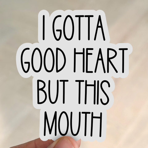 A hand holds a white sticker with black playful lettering that reads, “I Gotta Good Heart But This Mouth.”