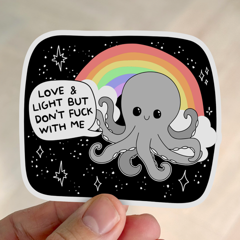 A hand holds a white sticker with black playful lettering that reads, “Love And Light But Don't Fuck With Me.”