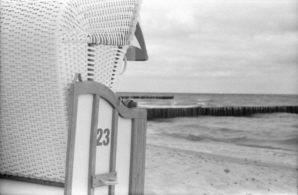 Eine schwarz-weiß-Fotorgrafie.
Das rechte Drittel ist ausgefüllt durch einen Strandkorb, von schräg hinten foografiert. An der Seite steht die Zahl 23. In Hintergrund Strand mit Buhnen, die ins Meer ragen.