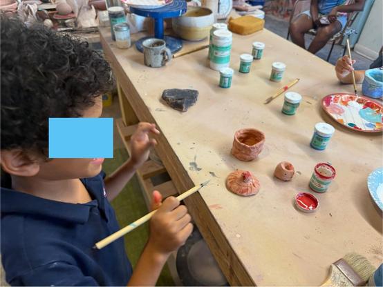 A little boy in a blue collared shirt with black curly hair holding a paint brush, standing at a table filled with pieces of pottery and little bottles of colored underglaze. In the upper right corner, you can see the hand of another child working across from him and the legs of one of the moms in the background. 