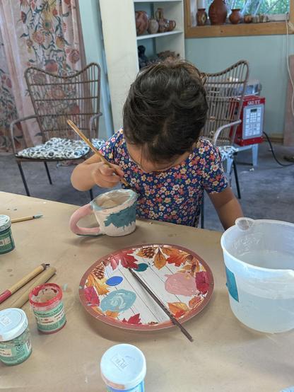 A little girl intently painting a ceramic mug. She is wearing a floral dress and the colors of her mug are aqua, white, light blue and pink. There is a paper plate on the table in front of her dotted with small amounts of different-colored underglaze. There is also a  large plastic measuring cup full of water, brushes and bottles of underglaze.