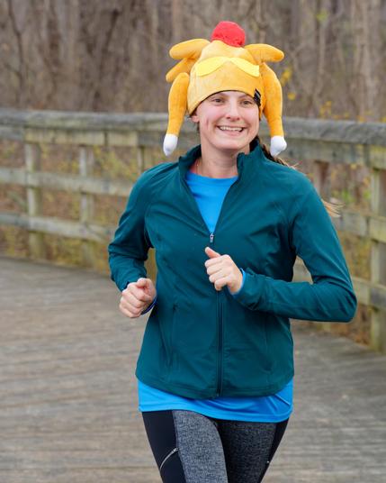 Woman in a blueish green jacket has a brighter blue shirt and black and grey pants and a big yellow, white and red turkey hat with a wood slat bridge in back