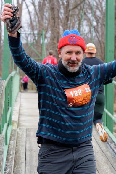 Man with red and blue hat and a dark blue shirt with pairs of horizontal light blue stripes and bib number 122 holds his hands up high at the close end of a pedestrial suspension bridge