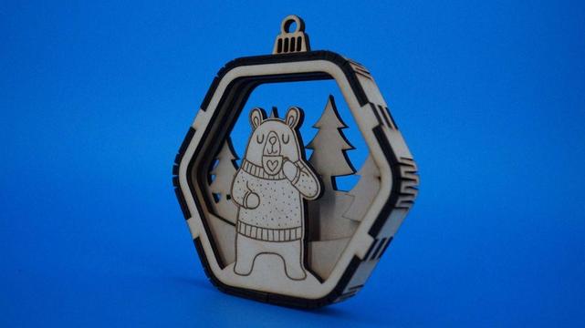 Holzanhänger in sechseckiger Kugelform, im Inneren steht ein gelaserter Bär mit Winterpullover und Herz-Tasse. Dahinter stehen ausgeschnittene Tannenbäume. Natürliche Holzfarben, feine Gravurdetails.