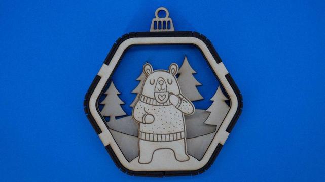Holzanhänger in sechseckiger Kugelform, im Inneren steht ein gelaserter Bär mit Winterpullover und Herz-Tasse. Dahinter stehen ausgeschnittene Tannenbäume. Natürliche Holzfarben, feine Gravurdetails.