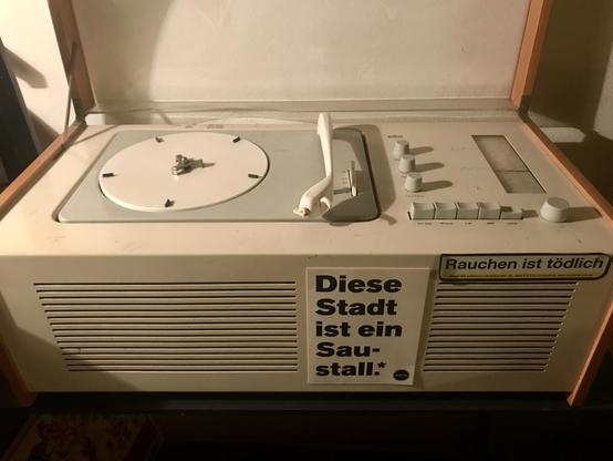 An old-fashioned record player with a wooden lid, featuring a circular turntable and several control knobs. It has a sticker in German that reads "Diese Stadt ist ein Saustall" (This city is a pigsty) and another saying "Rauchen ist tödlich." (Smoking is deadly)