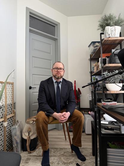 A man is sitting in a chair. He is overdressed for what his day will entail–sitting at home on video conferences all day. Background is a messy office.