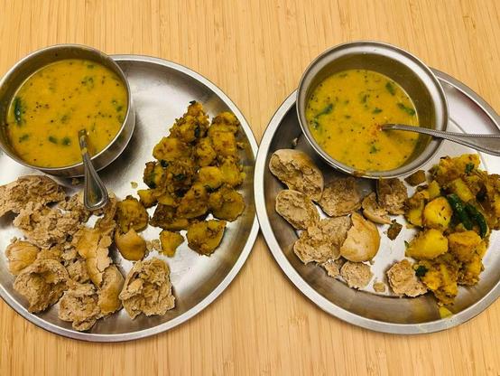 Two stainless steel plates with stainless steel bowls. The bowls have a yellow dal with coriander garnish. On the lower right side of both plates are crumbled wheat bati (dumplings) and next to them are spiced potatoes. 