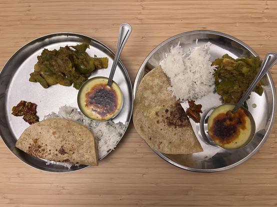 Two stainless steel plates with rice, roti, a red chili pickle. There is a potato and green pepper subzi and a small steel bowl with a yellow kadhi with a drizzle of red chilli tadka. 
