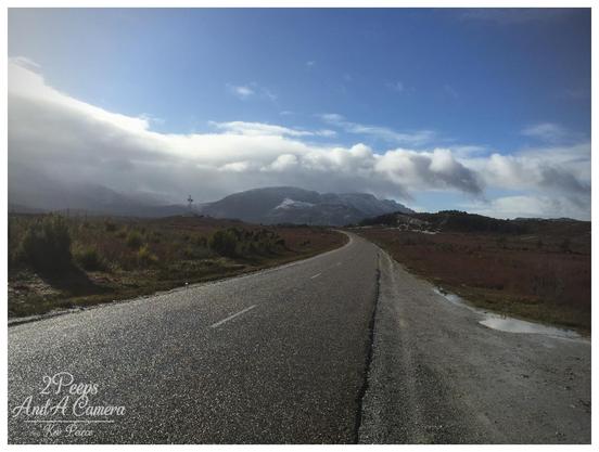 A moody photograph looking down a wet, winding asphalt road leading away from Strahan, Tasmania.

The roadside is covered in wet scrub and marshy ground, with distant hills shrouded in mist and snow dusted mountains beneath a dramatic sky where sunlight breaks through dark, rolling clouds.