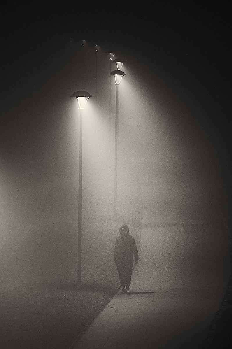 A lone figure in a dark hooded coat walks towards the camera along a narrow path at night, almost swallowed by the dense fog that softens every edge. Tall streetlamps stand in a diagonal line receding into the distance from the top left of the frame, each casting a narrow beam of warm light that pierces the mist and fades into the darkness. The person's face is hidden in shadow, their body only partially defined, adding to the sense of anonymity and unease. The grainy texture and monochrome tones make the scene feel both cinematic and isolated.