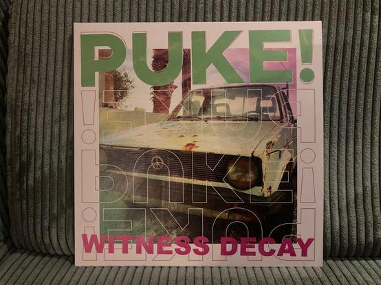 Das Bild zeigt ein Cover-Design mit dem Wort „PUKE!“ in auffälligen grünen und pinkfarbenen Buchstaben vor dem Hintergrund eines alten, rostigen Autos auf einem abgenutzten Foto. Der Titel „WITNESS DECAY“ ist in fetten pinkfarbenen Buchstaben am unteren Rand geschrieben.