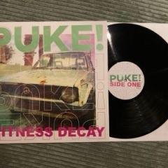 Eine Vinylplattenauslage mit einer schwarzen Platte neben ihrem Albumcover. Das Cover trägt den Titel „PUKE!“ und zeigt ein Bild eines Oldtimers mit dem auffällig platzierten Schriftzug „FITNESS DECAY“.