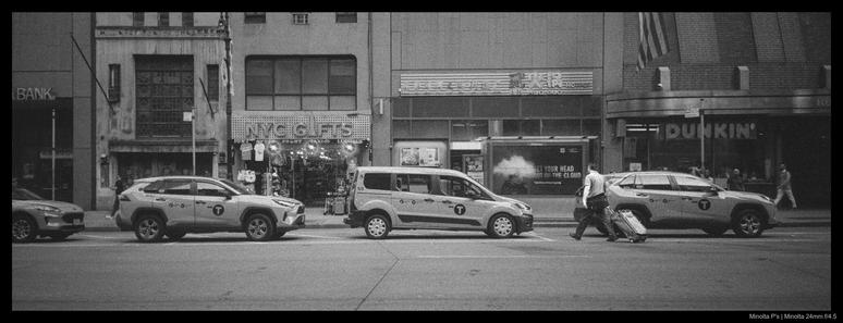 A person drags a suitcase behind them up to a line of waiting taxis, crossing the street outside of any crosswalk.  Shops are visible in the background behind the parked cars..
