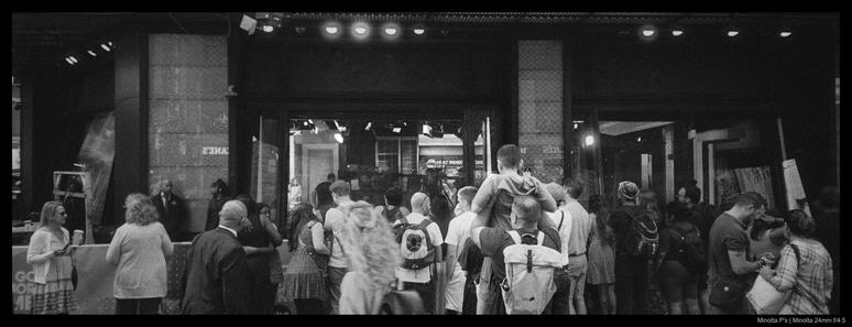 A small crowd of people gather outside the windows of Good Morning America's Times Square studio.  One child sits atop their parent's shoulders; the news desk and staff are visible through the large glass windows in front of them.