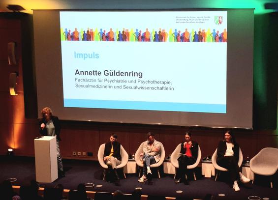 Podiumsdiskussion in einem Auditorium: Fünf Personen sitzen auf einer Bühne, eine Person steht am Rednerpult und spricht ins Mikrofon. Auf der großen Leinwand hinter ihnen ist eine Präsentationsfolie mit dem Titel „Impuls“ und dem Namen Annette Güldenring zu sehen, Fachärztin für Psychiatrie, Psychotherapie, Sexualmedizin und Sexualwissenschaft. Die Veranstaltung wird vom Ministerium für Kinder, Jugend, Familie, Gleichstellung, Flucht und Integration NRW ausgerichtet.