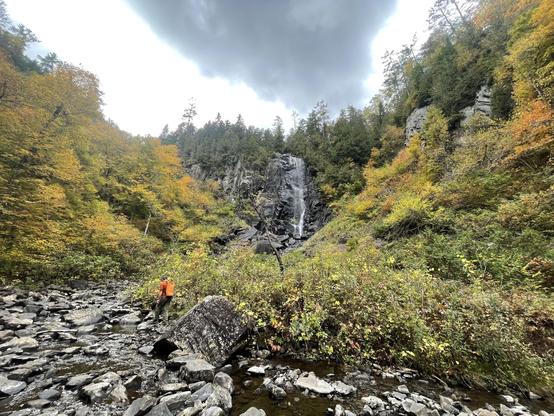 A rocky bluff with a tall waterfall. A hiker in an orange top is dwarfed as they ascend to the waterfall. Autumn trees line some of the rocks