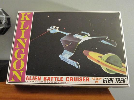 Box of a model kit. The 1/650 Klingon Battle Cruiser from AMT/Round2 Models. Back-left illustration of the ship in space near a ringed planet. Box is on a wooden shelf with a grey background.