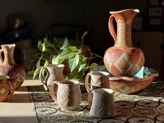 A collection of handcrafted pottery is displayed, including a large, intricately designed pitcher and several smaller mugs. The pottery features geometric patterns and earthy tones, set against a patterned cloth and accompanied by greenery. The late afternoon sun shines brightly on the pots with deep shadow in the background. 