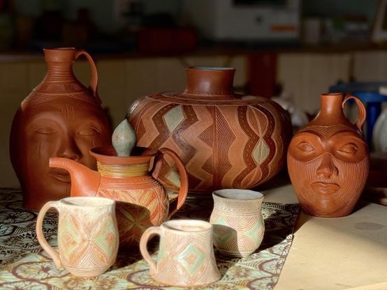 A collection of handcrafted pottery featuring various designs. The arrangement includes a large pot with geometric patterns, a teapot, and two mugs, alongside two vessels with face motifs. The pieces are made from terracotta and showcase intricate carvings and earthy tones. Blurred in the background are various office items in the back of my studio such as a printer. The pots are in the foreground beautifully lit by the sun. 