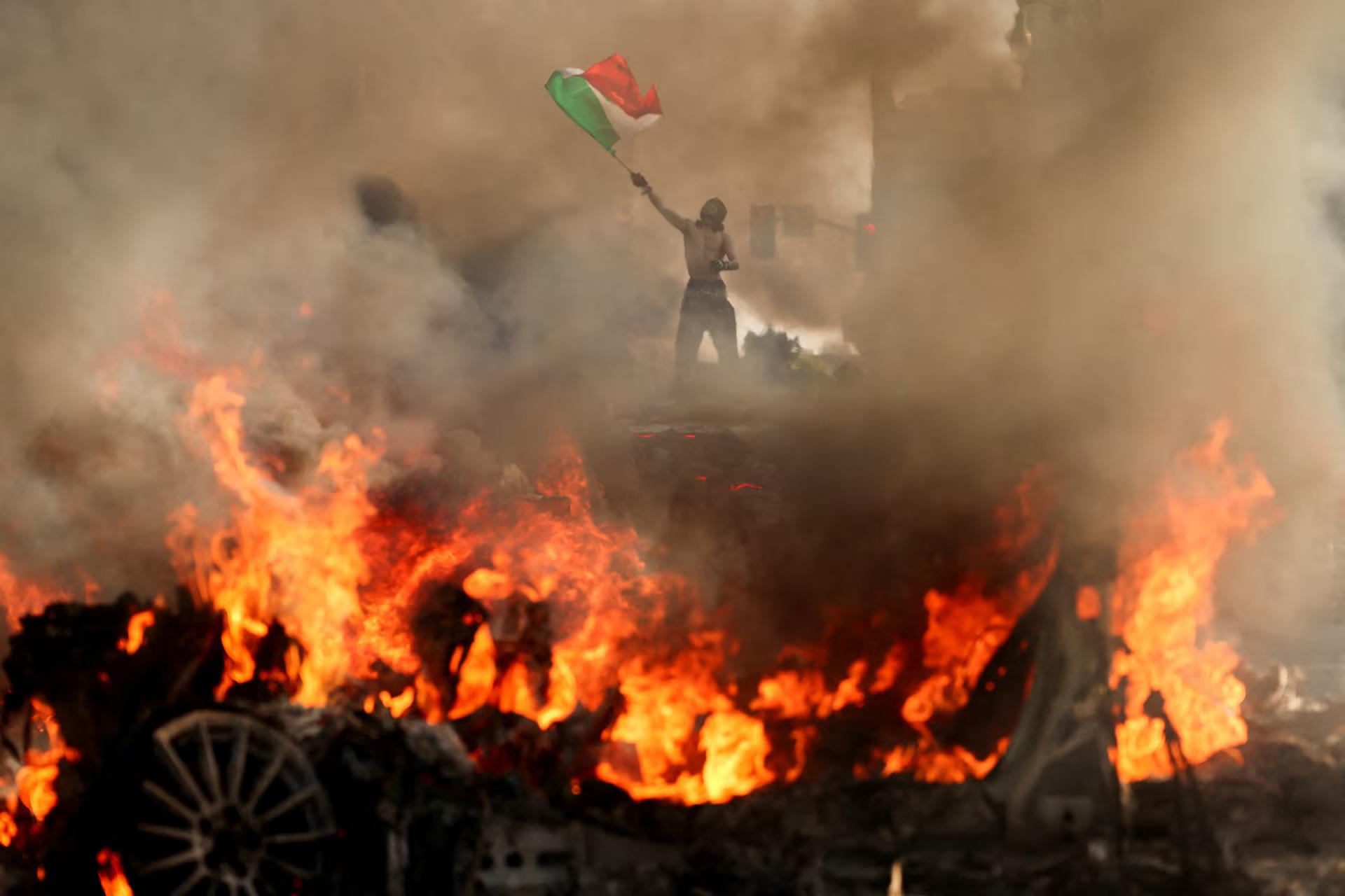 A man waves a Mexican flag as smoke and flames rise from a burning vehicle during a protest against federal immigration sweeps, near Los Angeles City Hall, in downtown Los Angeles.