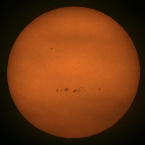 A close-up image of the sun, showing a bright orange surface with several dark sunspots visible. The background is dark, emphasizing the sun's glow.