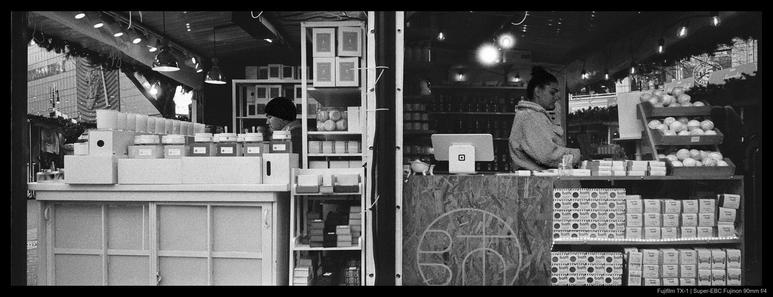 Two shopkeepers sit in their back-to-back stalls, shown from the sides, while selling their wares at the Union Square Holiday Market.  A large collection of goods are visible arranged on shelves in both shops.  Both people are looking down at something.