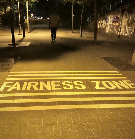 A dimly lit pathway at night featuring the phrase "FAIRNESS ZONE" painted on the ground. A person is walking in the distance, surrounded by trees and street lighting.