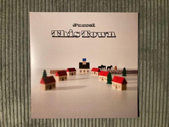 Das Bild zeigt ein Design für ein Musikalbumcover mit dem Titel „This Town“ des Künstlers Jussel. Es stellt eine Miniaturstadt mit kleinen Holzhäusern, Bäumen und einigen Figuren vor einem weißen Hintergrund dar.