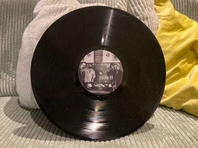 Eine schwarze Vinylplatte liegt auf einem grünen Stofftuch, in deren Mitte ein Etikett mit der Aufschrift „Side B“ und einem Schwarz-Weiß-Bild zu sehen ist. Weiche Kissen im Hintergrund verleihen der Szene zusätzliche Struktur.