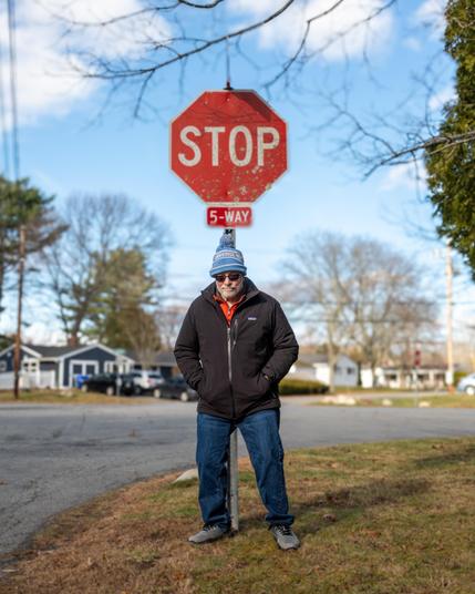 Color photo of a man bundled up for the cold standing just in front of of a stop sign on a sunny day.
