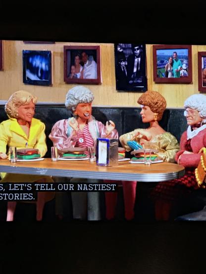 A group of four dolls resembling older women sit at a table in a diner, each holding a plate of food. They are engaged in conversation, with colorful outfits and distinctive hairstyles. The background features framed photos on the wall.