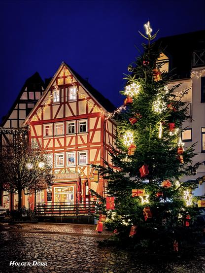 Abendliche Altstadt von Laubach im Advent: Ein hell geschmückter Weihnachtsbaum mit leuchtenden Sternen und roten Geschenkpaketen steht auf nassem Kopfsteinpflaster. Dahinter erhebt sich ein warm beleuchtetes Fachwerkhaus, dessen Lichterketten im tiefblauen Himmel schimmern. Die Szene wirkt festlich, ruhig und einladend – ein Moment voller vorweihnachtlicher Wärme.