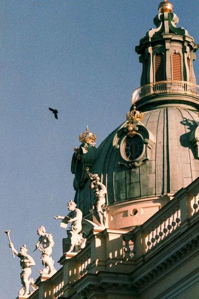 Partial view of a baroque building dome with gilded crowns and statues, featuring a bird in flight against a clear blue sky.

