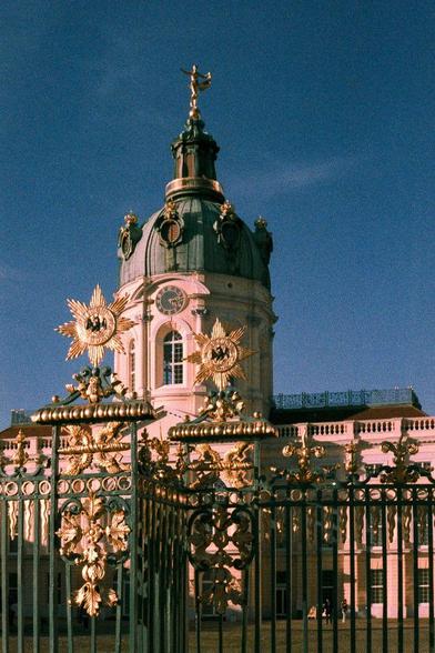 
View of Charlottenburg Palace in Berlin, Germany, behind a decorative gold and green gate. The palace features a large domed roof topped with a golden statue, a clock face, and two golden starburst decorations on the facade. The sky is a clear blue.

