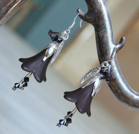 Black flower earring with silver petals, leaf and earring hooks. 3 small black crystals hand beneath the large tulip shaped flower. Both hang by their hoods from an vintage candelstick, blurry room behind.