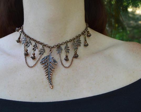 Copper fern leaf choker, worn by a pale figure in a forest. A large central fern leaf hangs from a chain, with smaller leaves either side. Bronze beads and loops of smaller chains hang from these leaves.