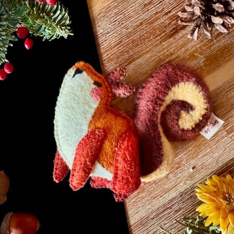 A small squirrel felt plushie standing on a wooden and black background, surrounded with decorative plants and acorns. The squirrel has a bright orange body and ivory-coloured belly. Its legs are a dark red and its twirly tail is brown and cream coloured, twirling around behind its back. Two small brown ears peak out of its head.  Embroidered on it’s face is a small squinty eye as well as a small heart-shaped nose and a pink blush.