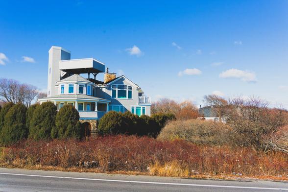 A house with observation deck fills about 1/4 of the area,  below is a clump of evergreen trees and some subtle colored winter phase shrubs with blue sky above with just a few clouds