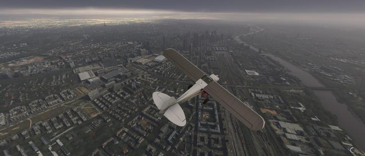 The image depicts a scene from a flight simulation, likely from a flight simulation software such as Microsoft Flight Simulator. The aircraft in the image is a biplane, which is a type of aircraft with two sets of wings, one above the other. The biplane is flying over a detailed urban landscape, which includes a mix of residential and commercial buildings, a river, and various roads and railways. The sky appears overcast, with a grayish tone, suggesting cloudy weather conditions. The overall scene is quite realistic, with attention to detail in the cityscape and the aircraft's design. The image captures the perspective of the pilot or the person flying the aircraft, providing a view of the city below and the surrounding environment.