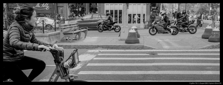 A bicyclist at a crosswalk begins to lurch into motion at the light.  In the opposite direction, a pack of motorcyclists are already crossing through the intersection.  Restaurants are visible in the background.