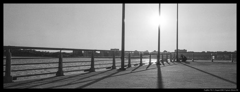 A person works on their bicycle by a set of railings overlooking the Hudson River.  Three tall poles throw long shadows from the sun, which hangs almost directly behind them in the sky.