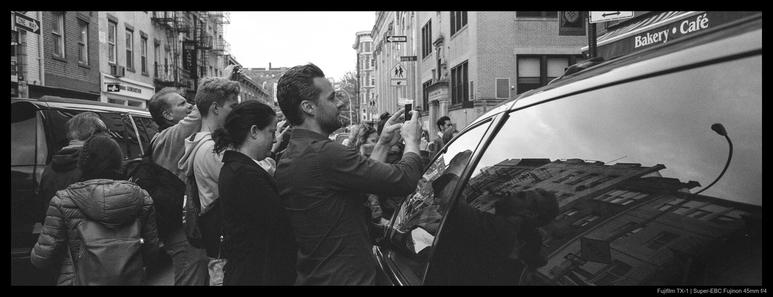 A large crowd of people spill into the street and ring a car as they try to record a curbside performance, itself out of view behind a parked car.