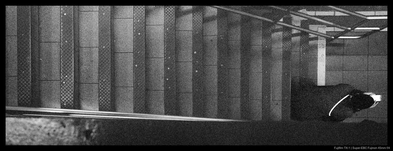 A person sits looking at their phone at the bottom of a flight of steps in the New York City subway system.  The photo was taken from above such that the stairs progress downwards from left to right.