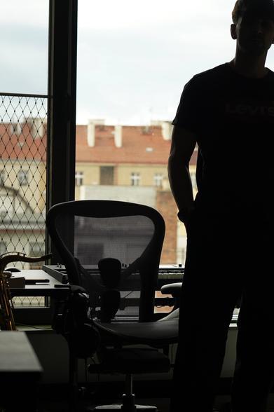 A photo of a man (myself) in front of a big window in a home office settings. There figure lacks colors (it is in the shade) due to a high contrast between the window and the lack of lighting indoors. The face is barely visible.