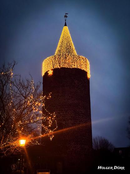 Der historische Diebsturm in Grünberg bei Abenddämmerung. Die Turmspitze ist vollständig mit goldenen Lichterketten geschmückt und leuchtet warm vor dem dunklen Himmel. Im Vordergrund stehen winterliche Äste mit kleinen Lichtpunkten und eine Laterne, die weiches, warmes Licht verbreitet. Das Bild wirkt feierlich, märchenhaft und winterlich.