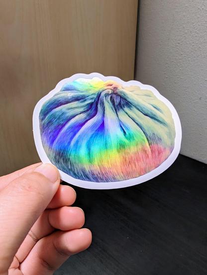 Photo of a left hand holding a diecut sticker. The sticker is a holographic dumpling drawing.