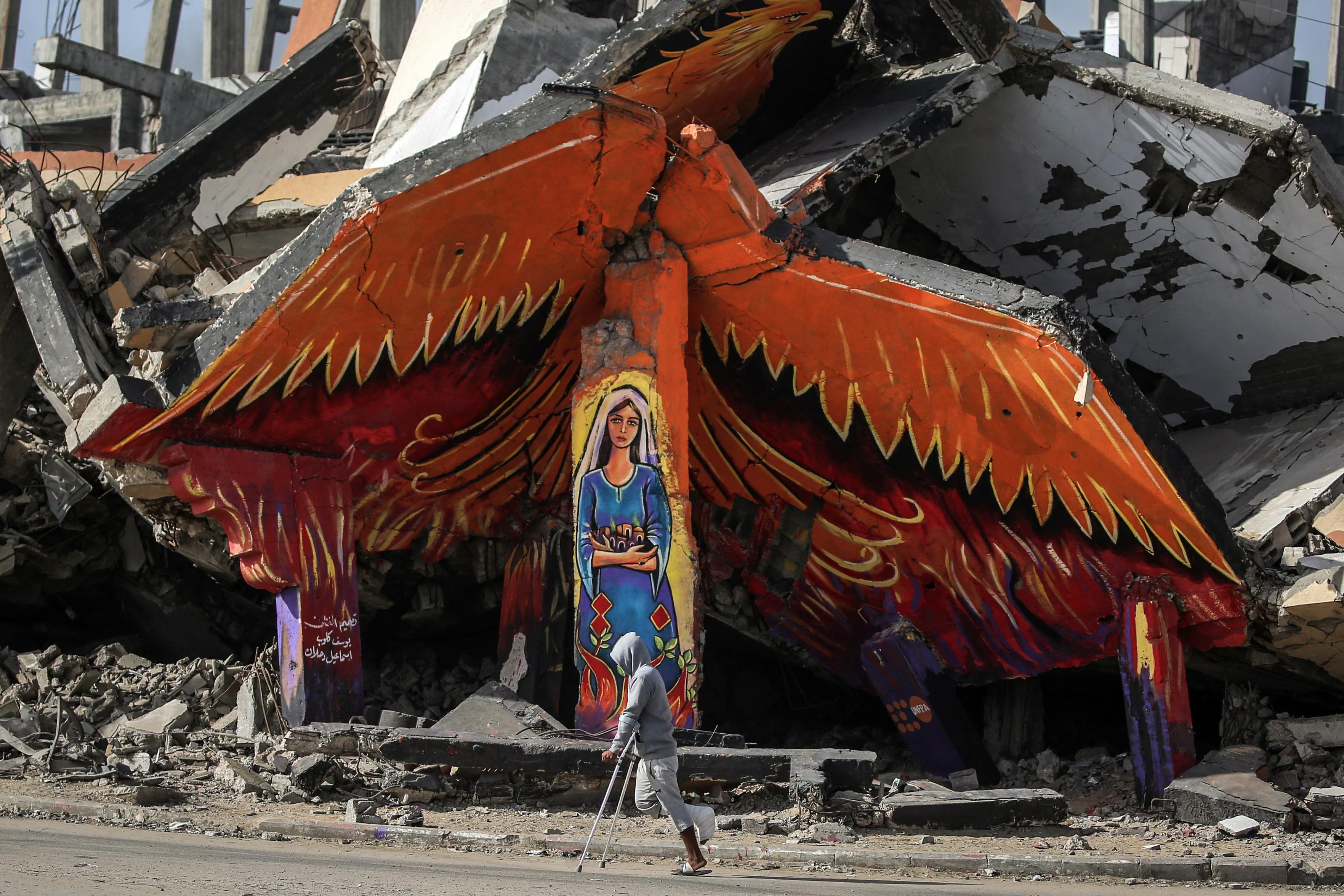 A man on crutches passes a mural of a Palestinian woman in a traditional dress on the side of a destroyed building in the Maghazi refugee camp.